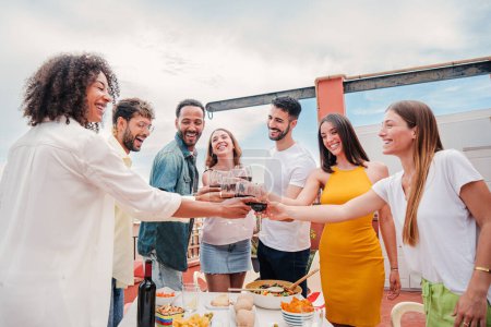 Group of multiracial adult friends smiling and clinking glasses of wine on a rooftop dinner party. Young happy people having fun toasting with alcohol celebrating together on a friendly meeting. High