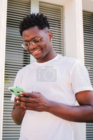 Téléchargez les photos : Portrait vertical de souriant jeune homme afro-américain utilisant son smartphone pour la messagerie dans les médias sociaux, regarder des nouvelles drôles. Beau mâle se connectant ou naviguant sur Internet avec un téléphone mobile - en image libre de droit