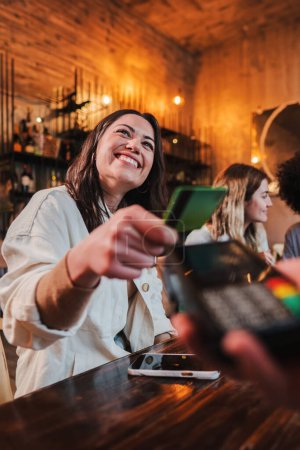 Photo for Vertical portrait of happy young woman paying bill with a contactless credit card in a restaurant. Cheerful female smiling holding a creditcard and giving a payment transaction to the cashier in a bar - Royalty Free Image