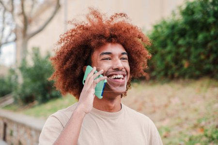 Téléchargez les photos : Jeune homme afro-américain souriant ayant un appel téléphonique mobile à l'extérieur. Adolescent heureux avec le sourire de dent parlant par téléphone portable. Jeune homme riant sur une conversation amicale smartphone sur le campus - en image libre de droit