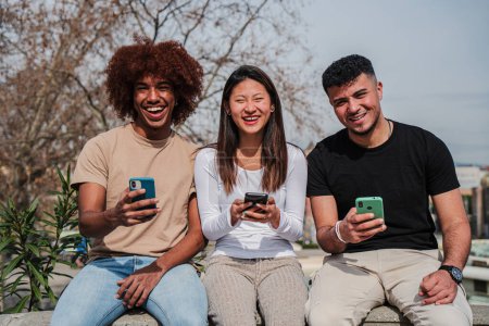 Téléchargez les photos : Groupe d'amis adolescents multiraciaux qui s'amusent à utiliser un appareil de téléphonie cellulaire à l'extérieur. Trois jeunes multiethniques souriants et riants regardant un partage de téléphone mobile sur les médias sociaux dans la rue - en image libre de droit