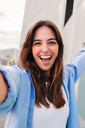 Téléchargez les photos : Portrait vertical d'une jeune étudiante caucasienne heureuse regardant la caméra et prenant un selfie s'amusant, debout à l'extérieur. Femme riante photographiant pour les médias sociaux sur le campus de l'université - en image libre de droit