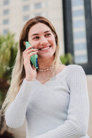 Foto de Retrato vertical de una joven joven bonita sonriendo y hablando con un dispositivo de teléfono inteligente. Mujer rubia feliz teniendo una conversación telefónica móvil en la calle urbana de la ciudad. Chica usando el teléfono celular fuera - Imagen libre de derechos