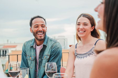 Photo for Smiling man laughing with two women friends celebrating a lunch party with wine glasses, sitting at table in the restaurant terrace. Group of three people relaxing and having a great time together - Royalty Free Image