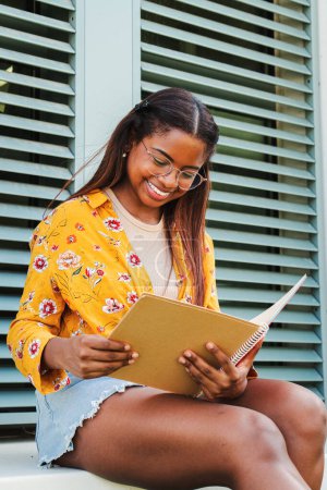 Foto de Retrato vertical de una joven latina feliz con gafas sosteniendo y leyendo un interesante libro en el campus universitario. Estudiante adolescente hispano estudiando la lección después de la clase. Concepto educativo - Imagen libre de derechos