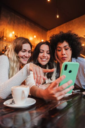Foto de Retrato vertical de un grupo de mujeres jóvenes divirtiéndose compartiendo medios con un teléfono celular. Mujeres mirando el teléfono inteligente en una cafetería. Chicas de compras en línea en un mercado usando un teléfono móvil - Imagen libre de derechos