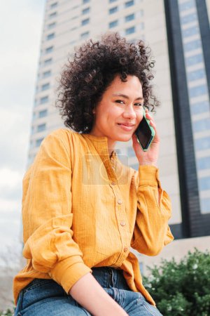 Téléchargez les photos : Portrait vertical de jeune femme latine aux cheveux bouclés ayant une conversation téléphonique avec un smartphone. Dame heureuse hispanique avec chemise jaune parlant par téléphone portable assis à l'extérieur. Femme utilisant le téléphone - en image libre de droit
