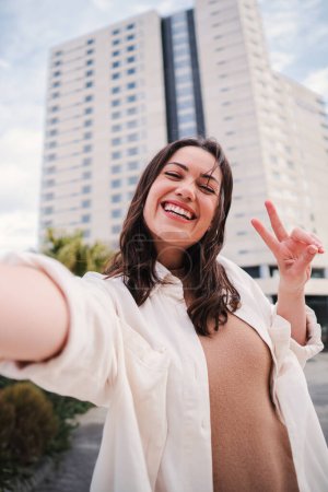 Photo for Vertical portrait of pretty young adult woman with thooty smile having fun taking a selfie using a social media. Happy teenage female laughing and looking at camera with cheerful expression outside - Royalty Free Image