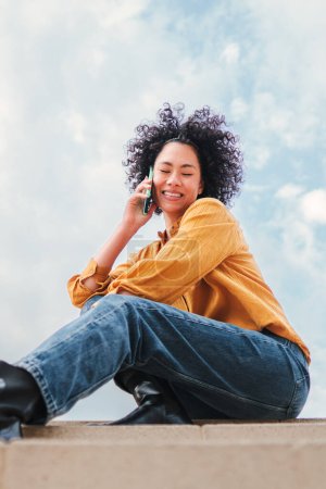 Téléchargez les photos : Portrait vertical de jeune femme hispanique adulte avec des cheveux bouclés ayant une conversation d'appel de téléphone portable assis sous le ciel bleu avec des nuages. Heureuse fille latine parlant par téléphone, à l'arrière-plan de la skyline - en image libre de droit