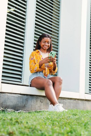 Téléchargez les photos : Portrait vertical d'une étudiante afro-américaine souriant à l'aide d'un téléphone portable, lisant des nouvelles impressionnantes ou faisant du shopping en ligne assis sur le campus universitaire. Joyeux adolescent femme regardant le téléphone mobile - en image libre de droit