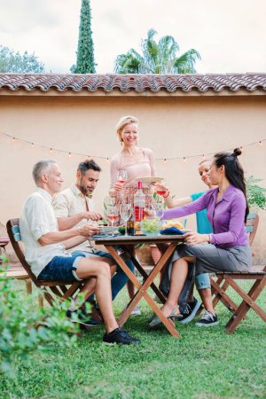 Téléchargez les photos : Portrait vertical d'un groupe d'amis adultes matures qui s'amusent ensemble lors d'un déjeuner barbecue à la maison. Personnes heureuses d'âge moyen souriant célébrant un anniversaire dîner réunion de famille - en image libre de droit