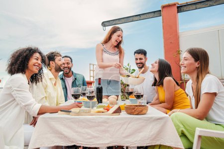 Foto de Mujer blanca alegre se reunió con sus amigos, sirviendo un almuerzo sentado a la mesa hablando y bebiendo vino, disfrutando de un gran tiempo juntos. Grupo de adultos jóvenes celebrando en la azotea de su casa - Imagen libre de derechos