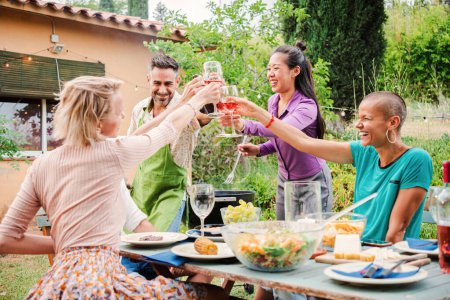 Téléchargez les photos : Des adultes enthousiastes levant leurs verres dans un toast joyeux lors d'une fête dans la cour pour célébrer l'amitié ensemble. Deux hommes d'âge moyen et trois femmes profitant d'un barbecue, cliquetis avec du vin - en image libre de droit