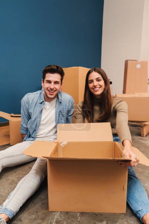 Foto de Vertical. Joven pareja caucásica sonriendo mudándose a su nuevo hogar, usando cajas de cartón para llevar sus cosas. Esposo y esposa sentados en el suelo de su apartamento desempacando cajas. Préstamo hipotecario - Imagen libre de derechos