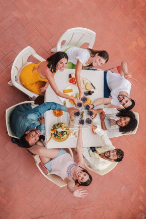 Photo for Vertical high angle top view of a group of best friends toasting wine glasses smiling and looking up to the camera. Young adult buddies people having fun on a dinner rooftop party clinking with drinks - Royalty Free Image