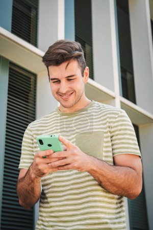 Téléchargez les photos : Portrait vertical d'un adolescent heureux souriant et tapant sur un téléphone intelligent à l'aide d'une application de médias sociaux à l'extérieur. Un jeune homme qui s'amuse à envoyer des messages sur son portable. Beau mec utilisant un téléphone portable - en image libre de droit
