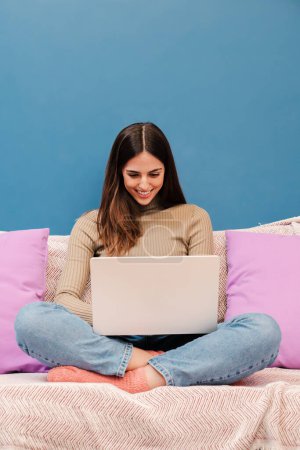 Foto de Retrato vertical de una joven que trabaja con una computadora portátil sentada en un sofá frente a una pared azul. Feliz hermosa mujer sonriendo usando un portátil con conexión inalámbrica a internet en casa. Alto. - Imagen libre de derechos