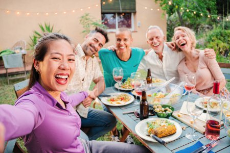Téléchargez les photos : Groupe d'amis adultes matures souriant prenant un selfie lors d'une fête barbecue à la maison arrière-cour. Les gens du Moyen Âge photographiant assis à la table de patio avec une expression positive et amicale - en image libre de droit