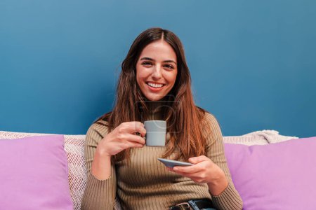 Photo for Portrait of caucasian young happy woman posing holding a coffee cup on her hand, smiling and enjoying the moment sitting on the sofa at her cosy apartment . Joyful female drinking a tea at home. High - Royalty Free Image
