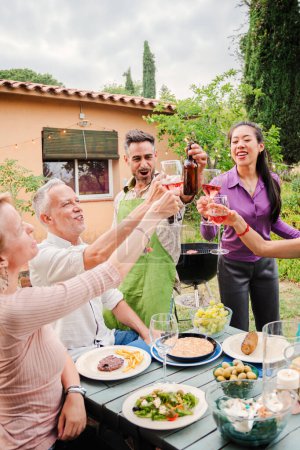Téléchargez les photos : Portrait vertical d'un groupe d'amis matures grillant des verres à vin rouge lors d'une fête de midi à la maison. Personnes heureuses d'âge moyen, boissons souriantes et clinquantes au barbecue réunion à l'extérieur - en image libre de droit