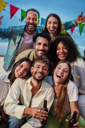 Vertical portrait of a big group of happy best friends smiling and having fun celebrating a buddies rooftop party, social gathering or birthday meeting at nightime, drinking beer and enjoying together