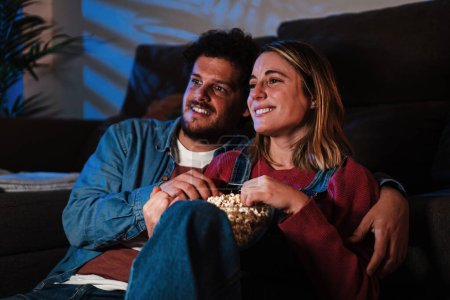Young caucasian couple smiling and having fun watching television show or movie at late night sitting on the sofa at home, eating popcorn. Two friends enjoying with streaming service on a smart tv