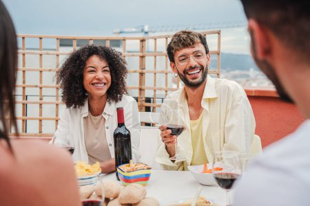 Téléchargez les photos : Rassemblement de gens profitant du beau temps assis à la table, parlant, buvant du vin à la fête sur le toit. Groupe souriant d'amis partageant de beaux moments ensemble, riant dans la célébration d'été - en image libre de droit