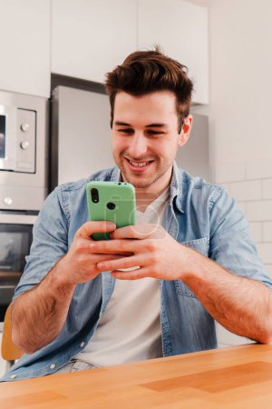 Téléchargez les photos : Portrait vertical d'un gars heureux utilisant son téléphone intelligent bavardant en envoyant des messages sur les médias sociaux assis à la cuisine de la maison et faisant une pause. Jeune homme souriant jouant à un jeu vidéo sur téléphone portable. Haut - en image libre de droit