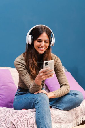 Foto de Retrato vertical de una joven feliz sonriendo y usando un teléfono inteligente para escuchar música con una aplicación impresionante y compartir fotos en redes sociales con teléfono celular y auriculares sentados en un sofá en casa - Imagen libre de derechos