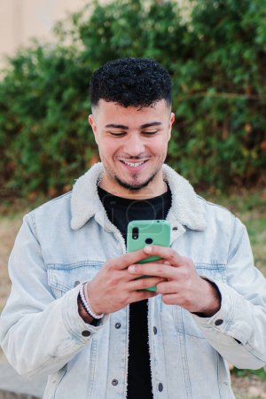 Téléchargez les photos : Portrait vertical d'un marocain souriant qui s'amuse à utiliser un smartphone pour jouer à un jeu vidéo. Heureux adolescents textos messages sur les médias sociaux avec un téléphone portable. Jeune homme avec un téléphone portable. Haut - en image libre de droit