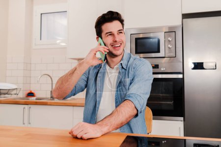 Téléchargez les photos : Jeune beau caucasien ayant une conversation téléphonique amicale assis à la maison avec une expression positive. Un jeune homme souriant, riant et parlant par téléphone portable dans la cuisine. Haut - en image libre de droit
