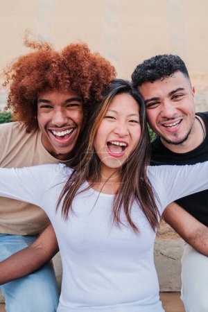 Foto de Retrato vertical de un grupo de adolescentes multirraciales riendo juntos. Mujer joven asiática extendiendo brazos y divirtiéndose con sus amigos masculinos. Gente feliz y emocionada vinculándose en una reunión - Imagen libre de derechos