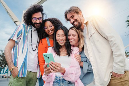 Photo for Multiracial group of young adult friends enjoying and smiling using a mobile phone app. Diverse buddies having fun watching and sharing content on the social media with a cellphone or smartphone. High - Royalty Free Image