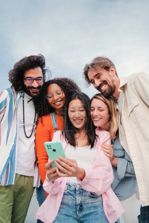 Photo for Vertical portrait of a multiracial group of young adult friends enjoying and smiling using a mobile phone app. Diverse buddies having fun watching and sharing on the social media with a cellphone - Royalty Free Image