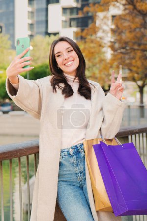 Téléchargez les photos : Portrait vertical d'une jeune femme heureuse souriante faisant un selfie après avoir fait du shopping et partagé sur les réseaux sociaux avec un téléphone portable. Une jolie fille isolée qui s'amuse à prendre une photo avec un téléphone portable - en image libre de droit