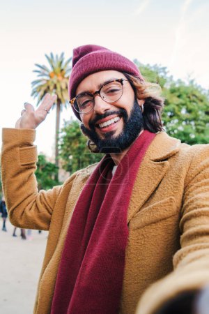 Photo for Vertical close up portrait of a happy man in a tourism trip. Young caucasian male taking a selfie with a smartphone in a park outdoors, wearing a beanie hat, glasses and scarf in autumn season. High - Royalty Free Image
