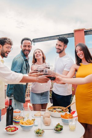 Téléchargez les photos : Portrait vertical d'un groupe d'amis adultes multiraciaux souriants et cliquetis verres de vin sur un dîner sur le toit. Jeunes gens heureux qui s'amusent à trinquer avec de l'alcool célébrant ensemble. Haut - en image libre de droit