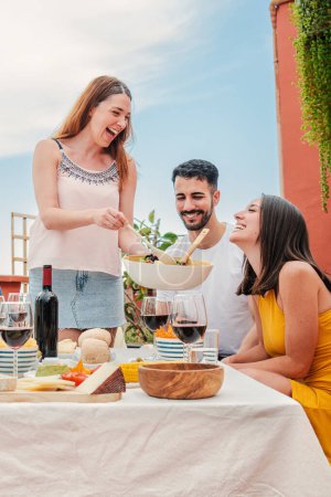 Téléchargez les photos : Portrait vertical d'une femme joyeuse réunie avec ses amis, servant un déjeuner assis à table parlant et buvant du vin, profitant d'un bon moment ensemble. Groupe de jeunes célébrant à la maison - en image libre de droit