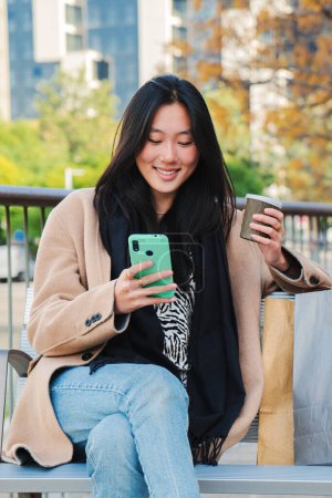 Téléchargez les photos : Portrait vertical d'une jeune femme asiatique insouciante tapant un message à l'aide d'un téléphone portable et prenant un café après les achats. Adolescent textos avec application smartphone assis sur un banc à l'extérieur. Communication - en image libre de droit