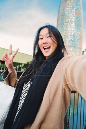 Foto de Vertical individual de cerca retrato de una joven asiática feliz tomando una foto selfie con un teléfono inteligente. Autorretrato de una mujer sonriente china haciendo el signo de paz después de ir de compras al aire libre. Alto. - Imagen libre de derechos