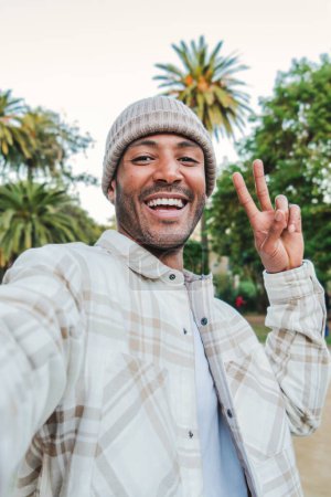 Téléchargez les photos : Portrait vertical individuel d'un jeune bel homme souriant faisant un portrait selfie avec un smartphone faisant le signe de la paix avec les doigts dans un parc à l'extérieur portant un bonnet. Concept de style de vie - en image libre de droit