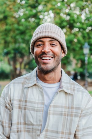 Photo for Vertical close up portrait of young hispanic man with a beanie hat smiling and looking at camera outdoors. Front view of latin happy guy standing at park with carefree attitude. Lifestyle concept - Royalty Free Image