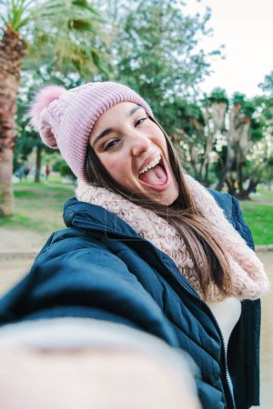 Foto de Retrato vertical de una joven mujer caucásica sonriendo tomando una selfie en un parque al aire libre con bufanda, gorro y ropa otoñal. Punto de vista de una adolescente haciendo una foto con un smartphone - Imagen libre de derechos
