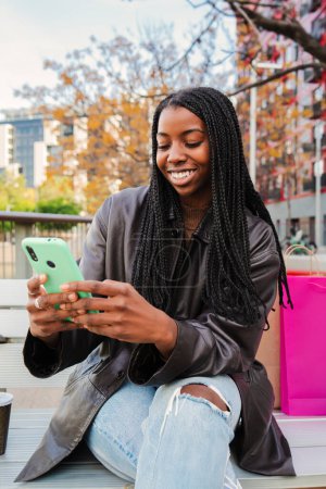 Téléchargez les photos : Portrait vertical d'une adolescente afro-américaine heureuse souriante faisant du shopping en ligne avec un téléphone portable assis sur un banc à l'extérieur. Jeune femme avec des tresses textos en utilisant un téléphone intelligent. Haute qualité - en image libre de droit