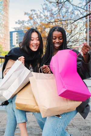 Foto de Retrato vertical de dos mujeres o niñas jóvenes multirraciales sonriendo y mirando dentro de una bolsa de compras sentadas en un banco. Un par de amigos felices mostrando sus compras en una semana de venta. Consumismo - Imagen libre de derechos