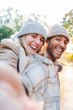 Photo for Vertical portrait of a young adult couple having fun together taking a selfie. Carefree boyfriend giving a piggyback ride to his cheerful girlfriend on a weekend sightseeing outside. High quality - Royalty Free Image