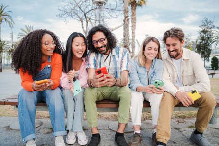 Téléchargez les photos : Groupe de jeunes amis adultes multiraciaux utilisant leur téléphone portable, naviguant sur Internet en profitant de temps libre assis sur un banc à l'extérieur. Rencontre de gens heureux s'amuser avec leurs téléphones intelligents - en image libre de droit