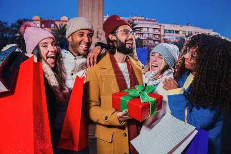 Photo for Group of multiracial young adult people giving a christmas present to their best friend. Happy man receiving a xmas surprise gift. Grateful guy holding a noel special wrapped box on a winter holidays - Royalty Free Image