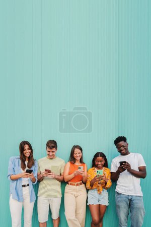 Foto de Vertical. Grupo multirracial de jóvenes amigos sonriendo con su aplicación de teléfono móvil en la pared verde azulado. Los adolescentes se divierten compartiendo mensajes en las redes sociales con un teléfono celular en un fondo de color azul. Alto. - Imagen libre de derechos