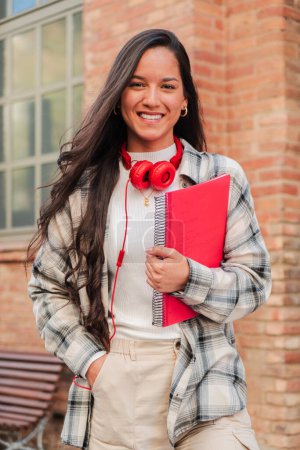 Téléchargez les photos : Portrait vertical individuel d'une jolie étudiante hispanique souriante et regardant la caméra au lycée. Plan de tête d'une adolescente latine debout à l'extérieur du campus universitaire. Éducation - en image libre de droit
